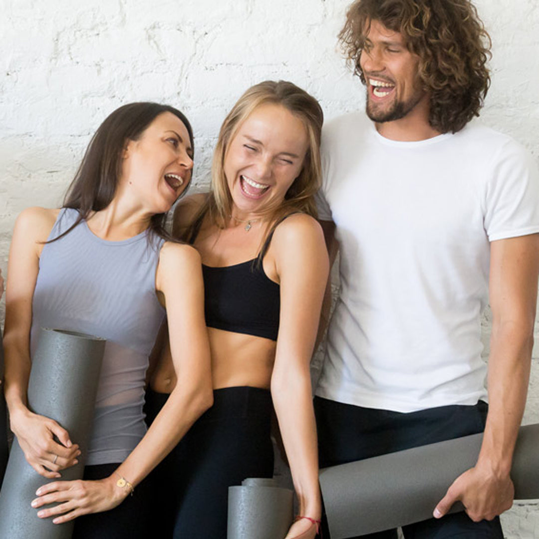 3 yogis (2 female + 1 male) standing together holding yoga mats, laughing together | TRIBE Yoga